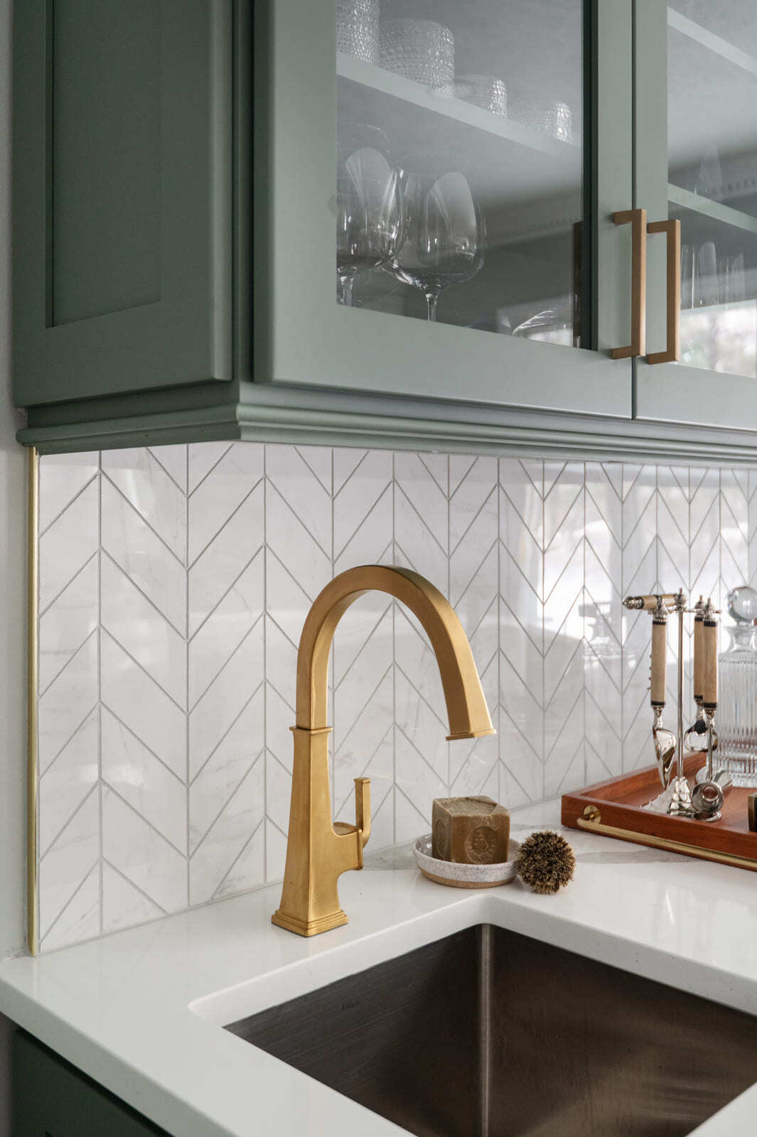 Brass faucet and bar sink beneath sage green glass-front cabinets with white chevron tile backsplash in Granger, IN