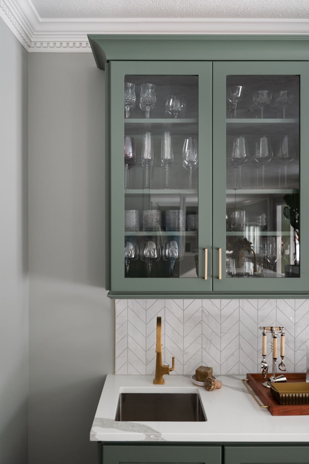Granger, IN kitchen bar sink with brass faucet, sage green glass-front cabinets, and white chevron tile backsplash