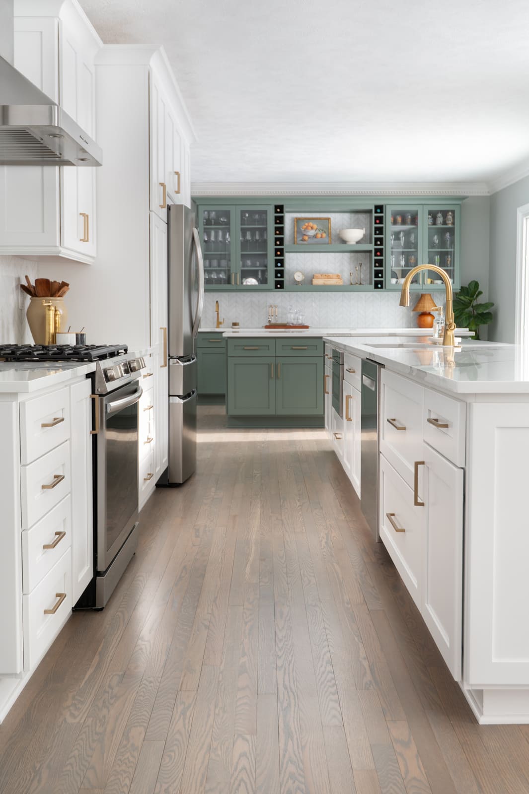 Long view of a Granger, IN kitchen with white cabinets, brass faucet, wood floors, and sage green bar in back