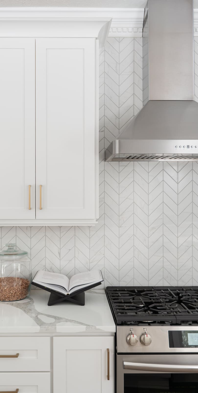 White kitchen cabinet and range area in Granger, IN with stainless hood, brass pulls, and chevron tile backsplash (1)