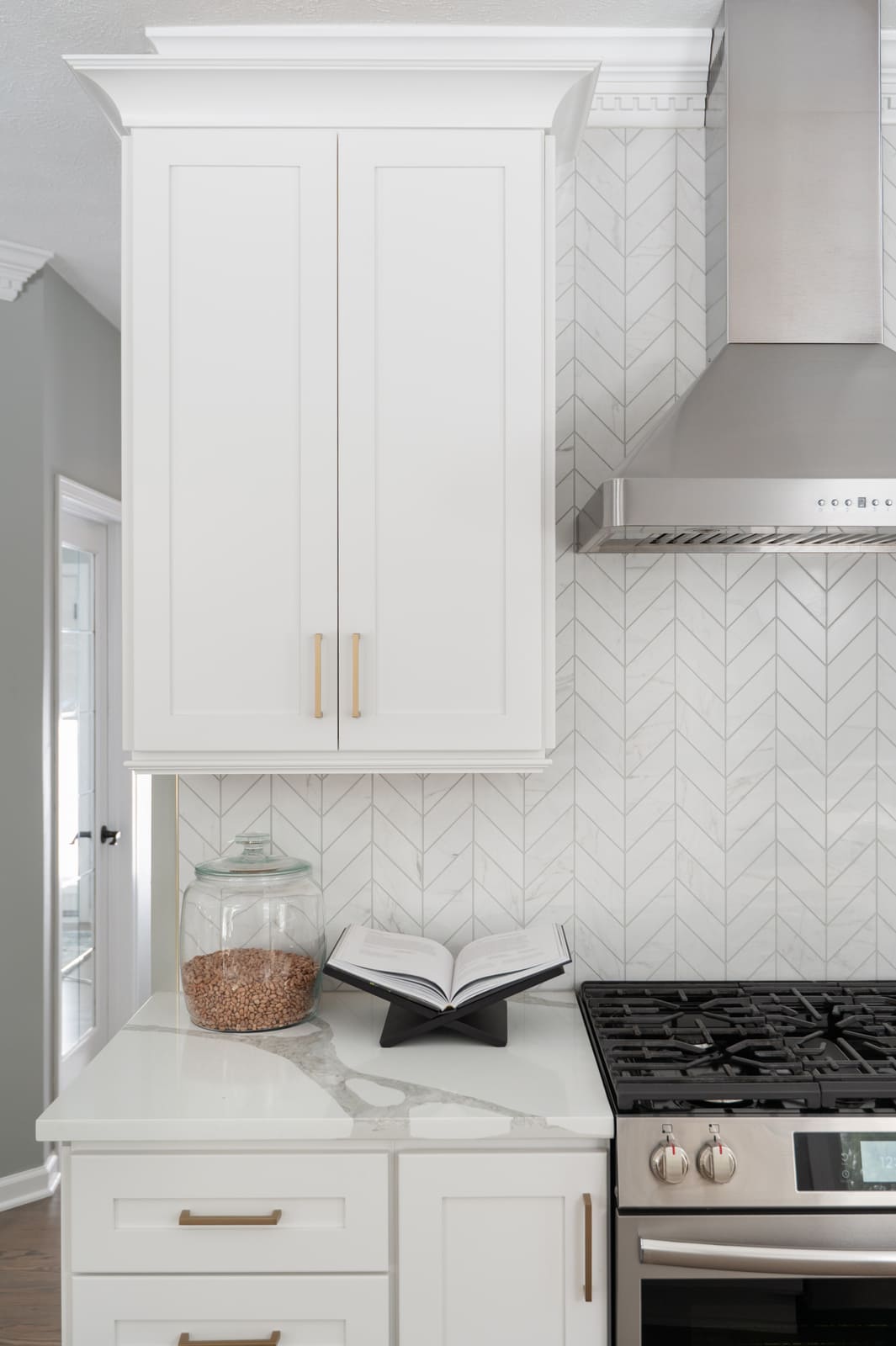 White kitchen cabinet and range area in Granger, IN with stainless hood, brass pulls, and chevron tile backsplash