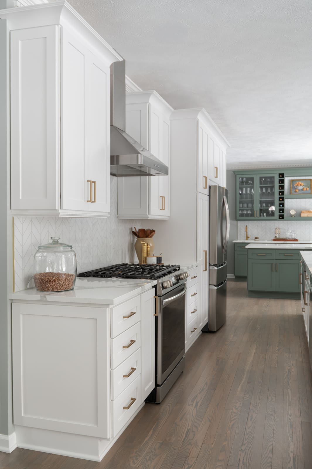 White kitchen in Granger, IN with stainless range hood, brass pulls, chevron tile backsplash, and view to sage green bar