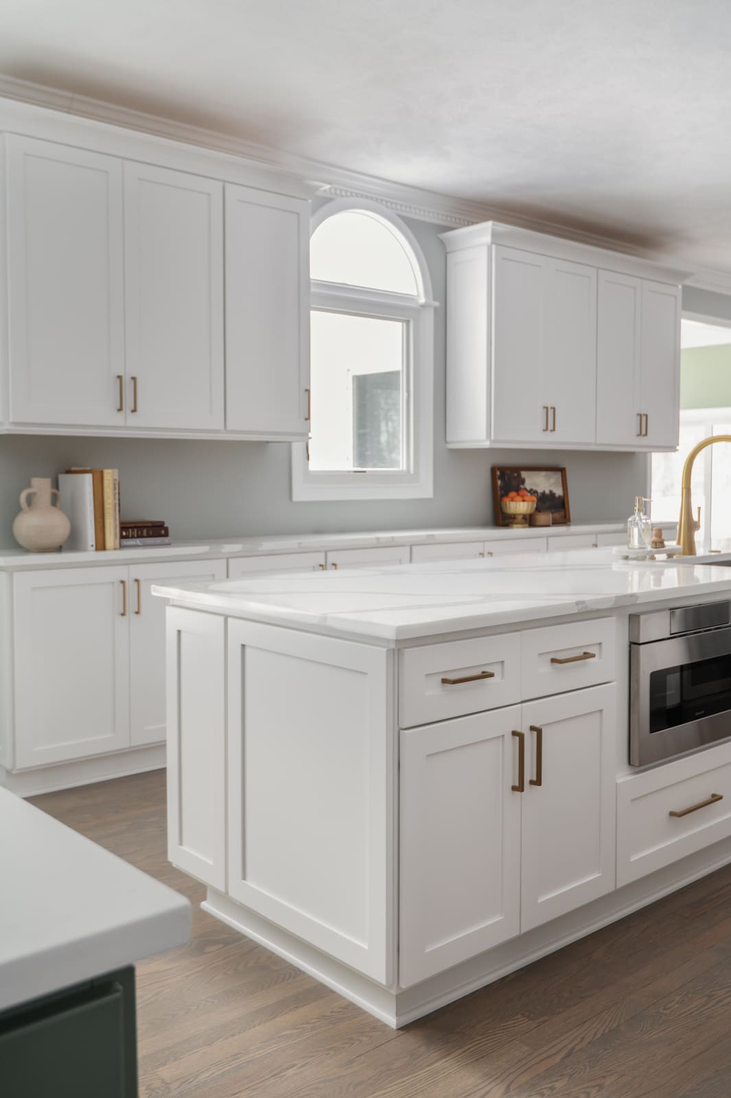 White kitchen island in Granger, IN with brass hardware, built-in microwave, and arched window between upper cabinets