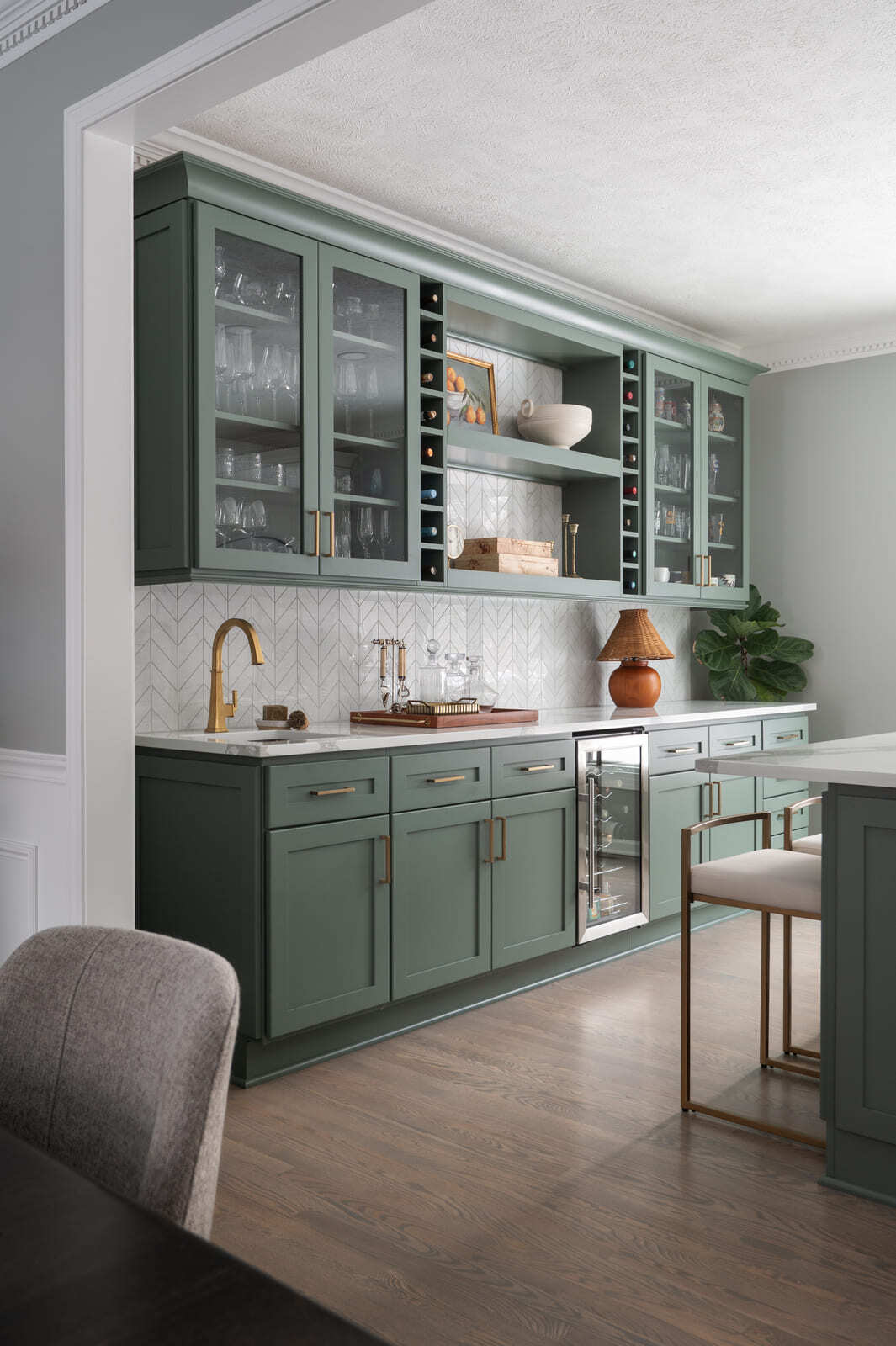 Wide view of sage green kitchen bar with wine fridge, glass cabinets, and brass accents in Granger, IN