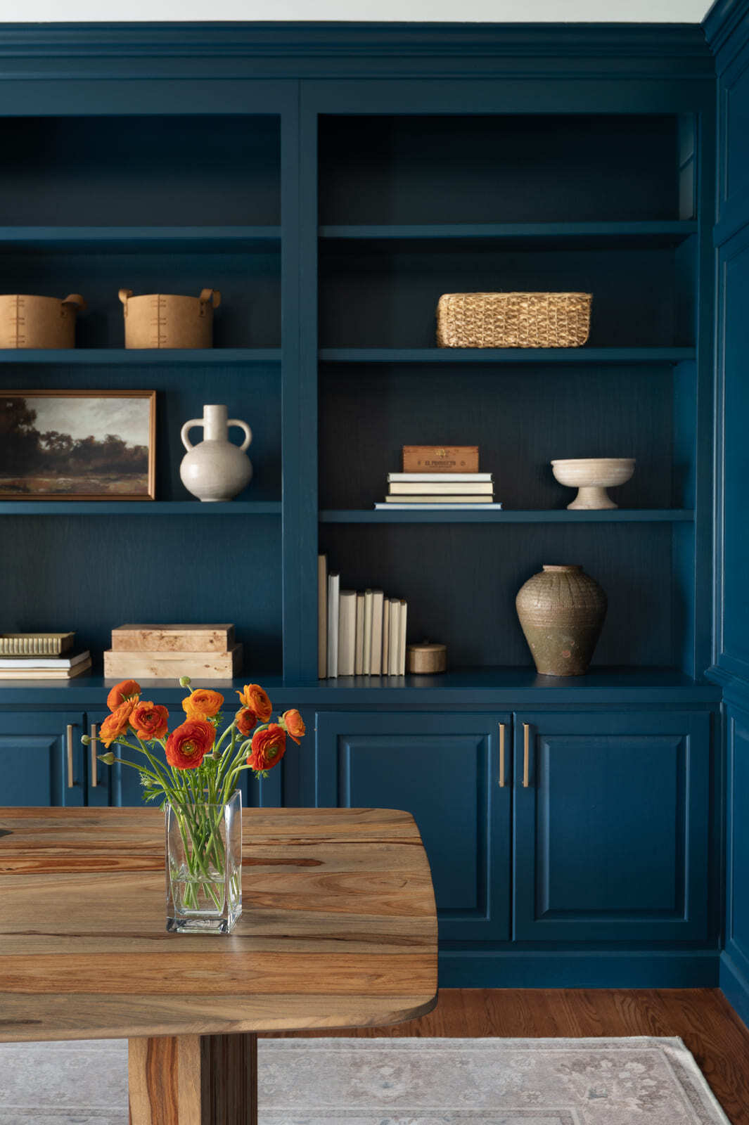 Blue built-in office shelves with books, baskets, pottery, brass hardware, wood desk, and orange flowers.