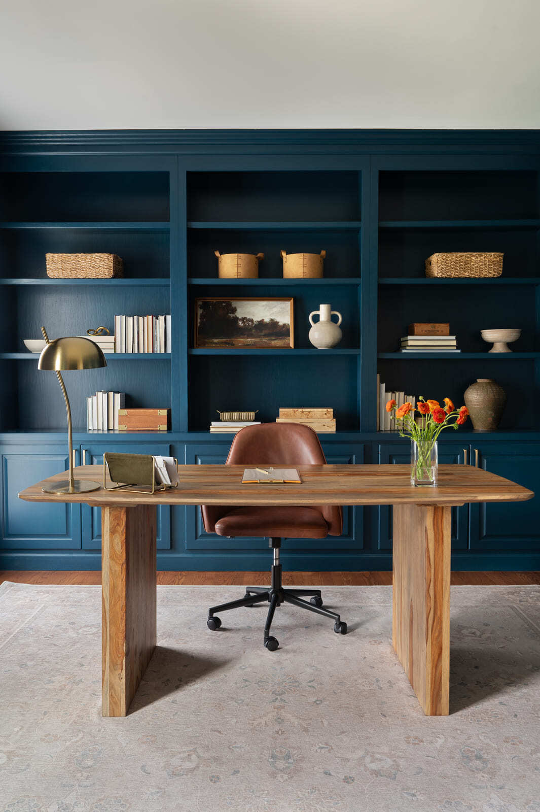 Blue home office with built-in bookshelves, wood desk, leather chair, brass lamp, area rug, and flowers.