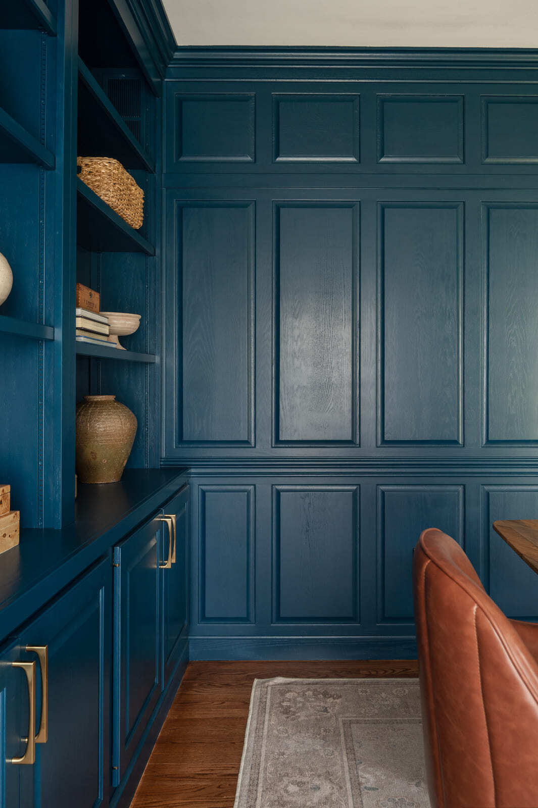 Blue home office with built-in shelves, paneled wall, brass hardware, leather chair, wood desk, and area rug.