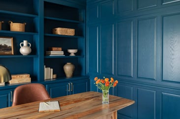 Blue home office with built-in shelves, paneled wall, wood desk, leather chair, orange flowers, and brass pulls.