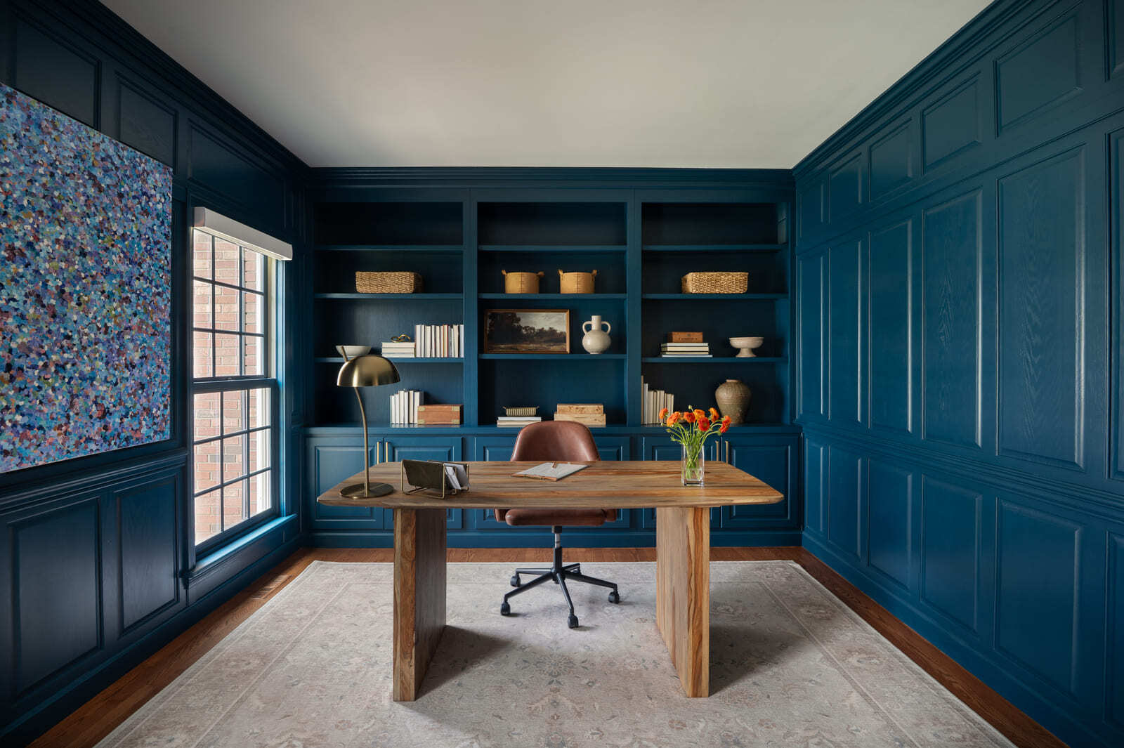 Blue home office with built-in shelves, paneled walls, wood desk, leather chair, window, abstract art, and area rug.