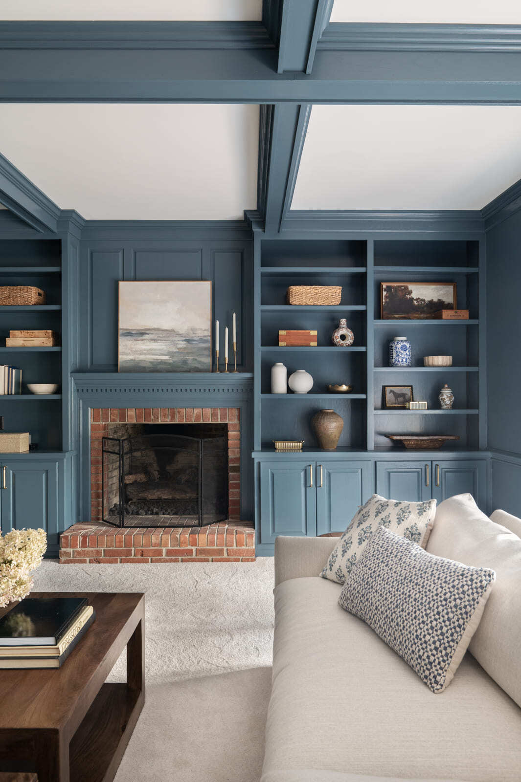Blue living room with coffered ceiling, brick fireplace, built-in shelves, white sofa, and patterned pillows.