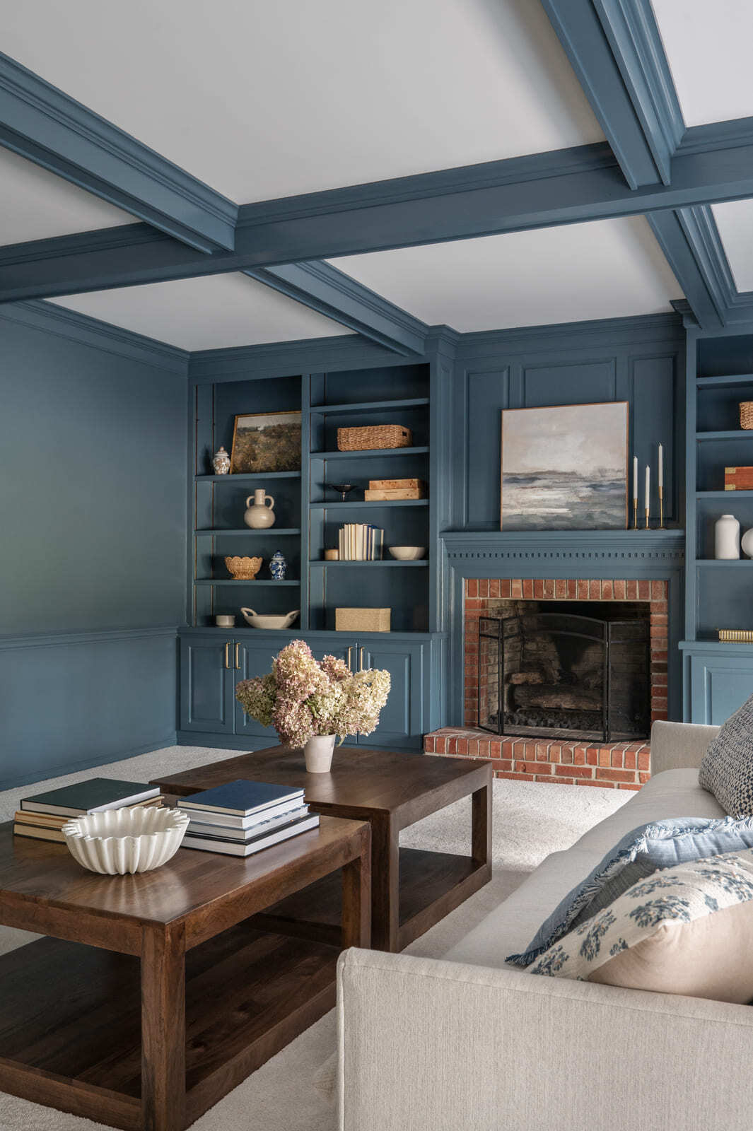 Blue living room with coffered ceiling, brick fireplace, built-in shelves, white sofa, and wood coffee table.