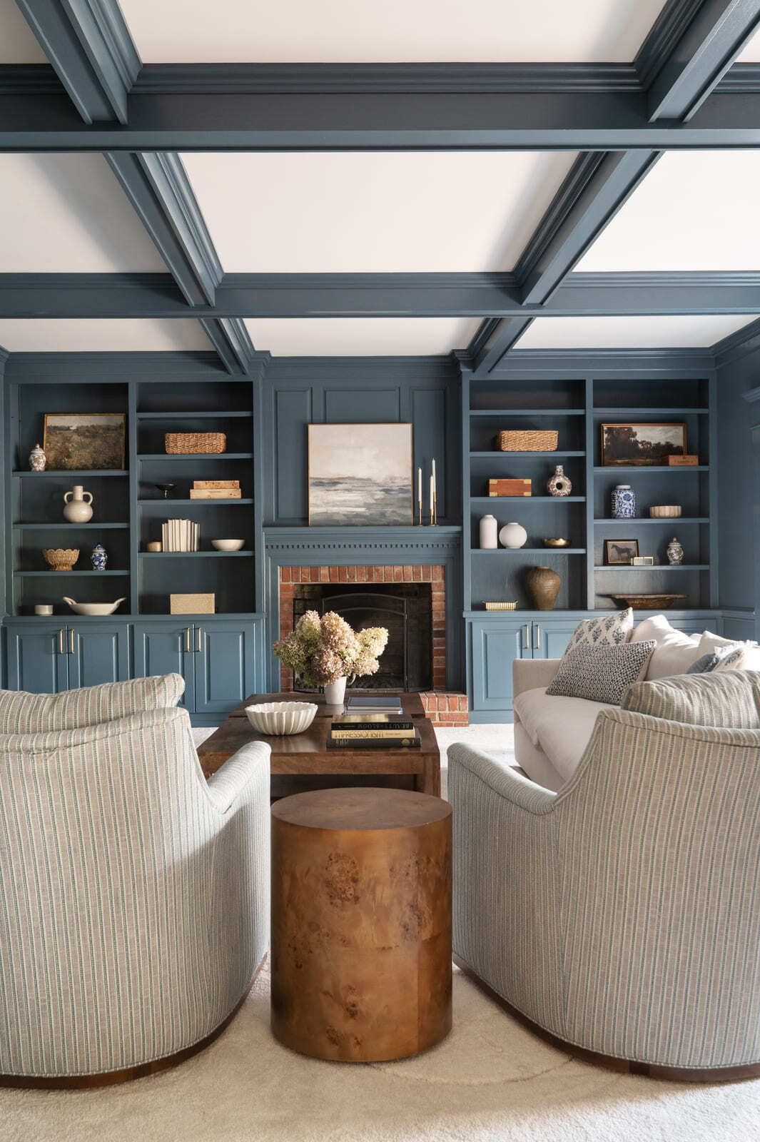 Blue living room with coffered ceiling, built-in shelves, brick fireplace, white sofa, chairs, and wood tables.