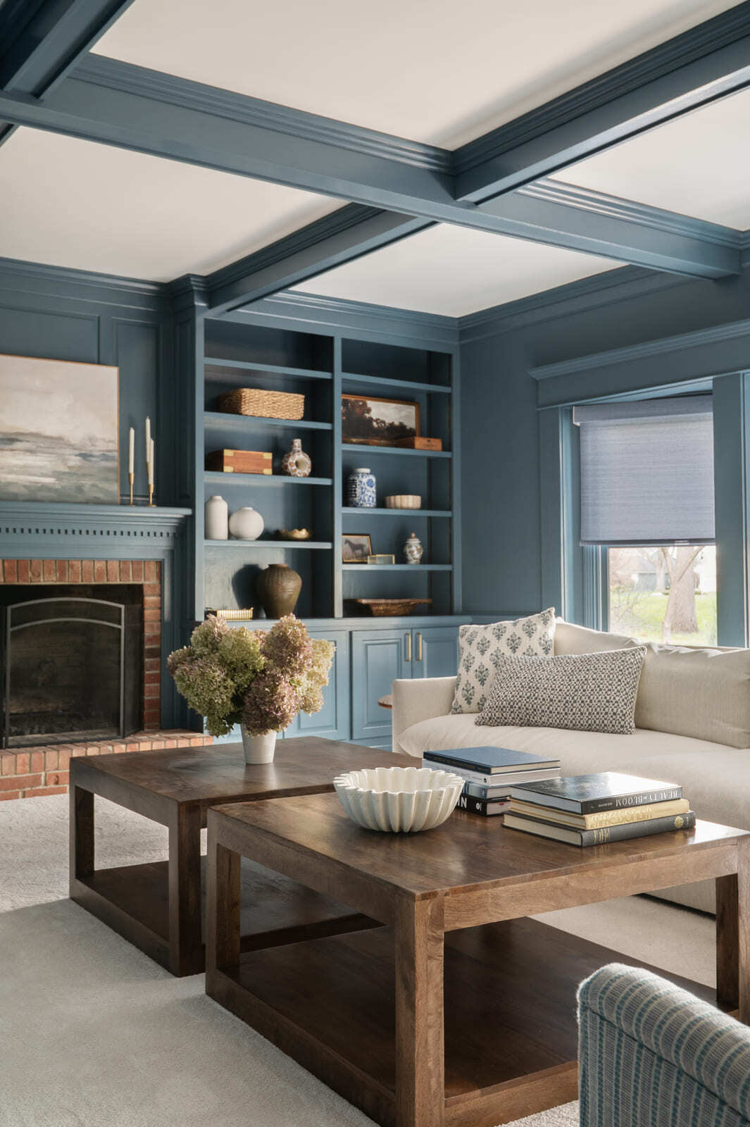 Blue living room with fireplace, built-in shelves, coffered ceiling, white sofa, wood coffee tables, and soft rug.