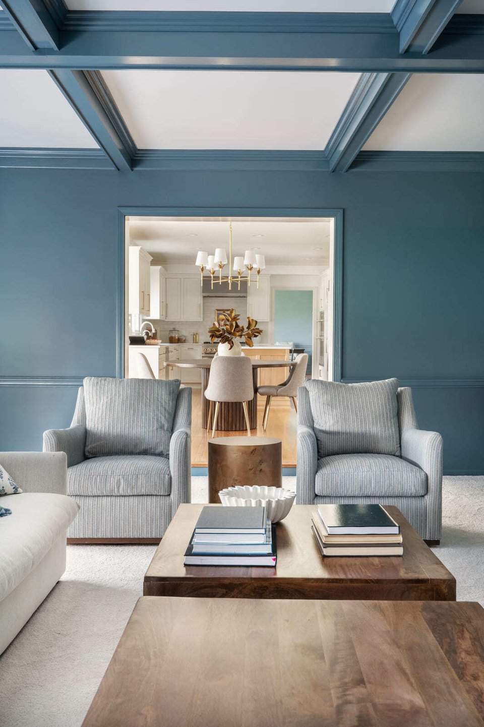 Blue living room with striped chairs, wood coffee table, coffered ceiling, and view into bright kitchen.
