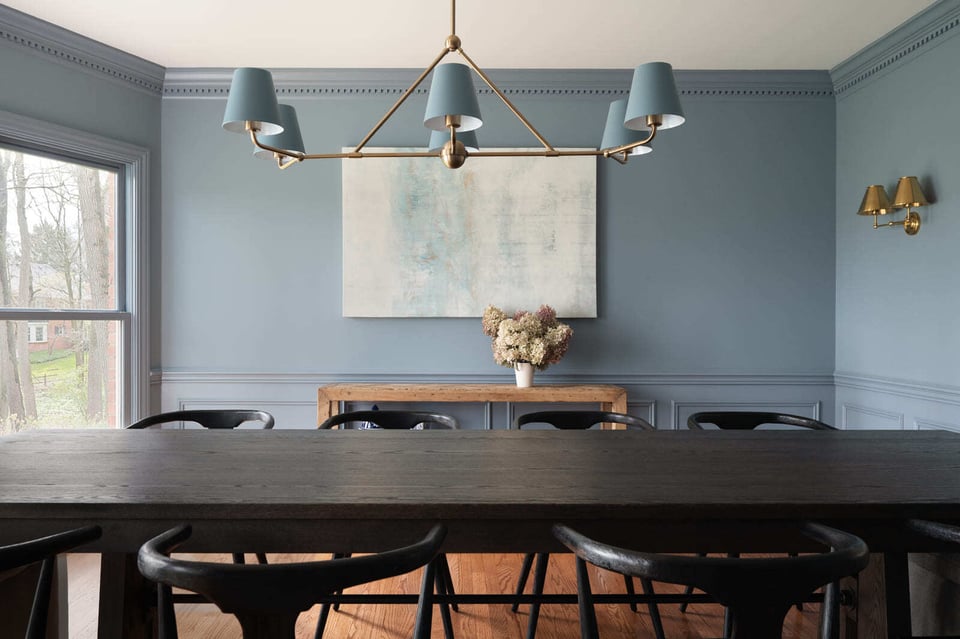 Bright blue dining room with large window, brass chandelier, dark wood table, black chairs, and abstract art.