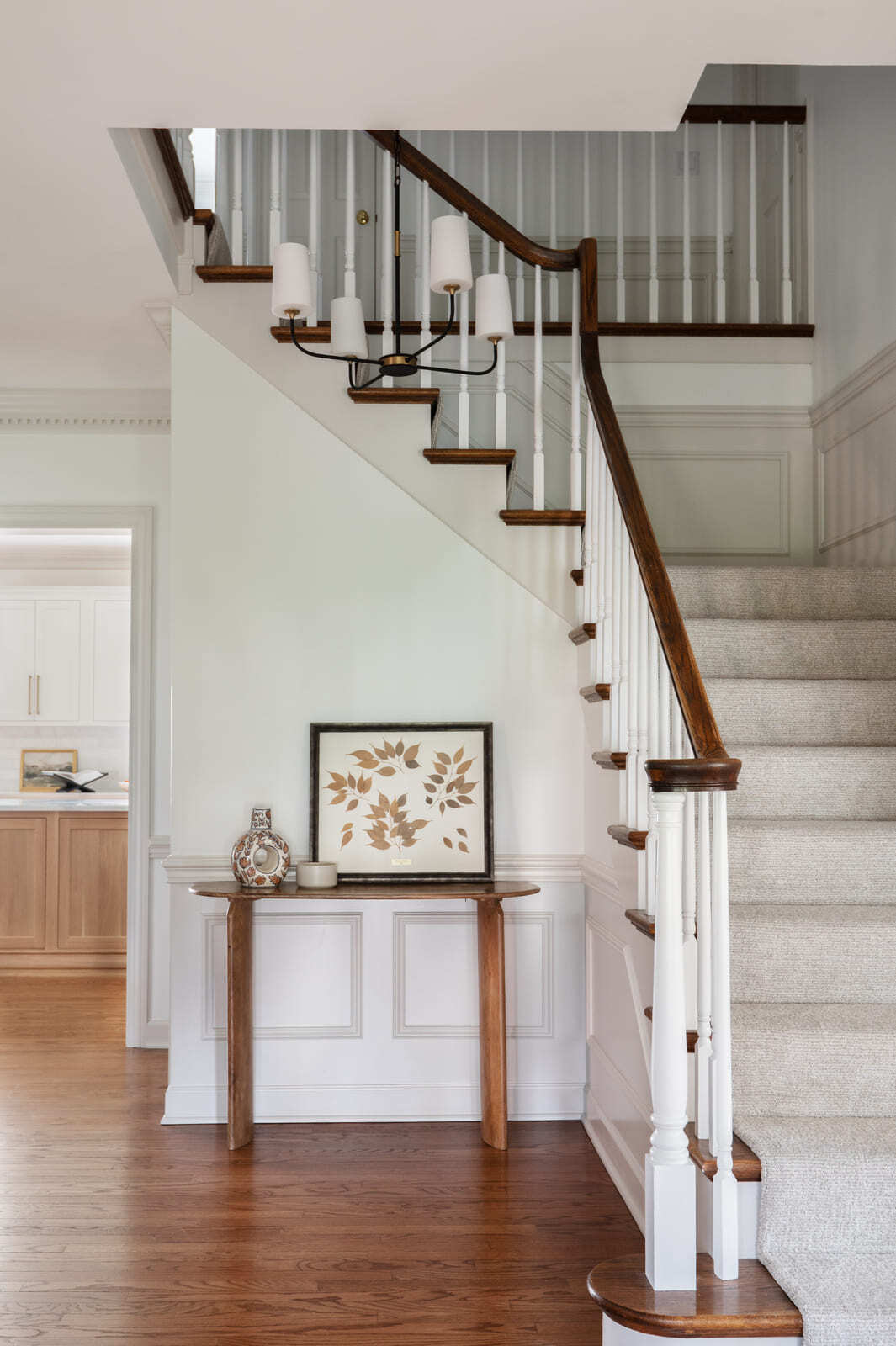 Bright foyer staircase with white spindles, wood handrail, carpet runner, console table, and kitchen view.