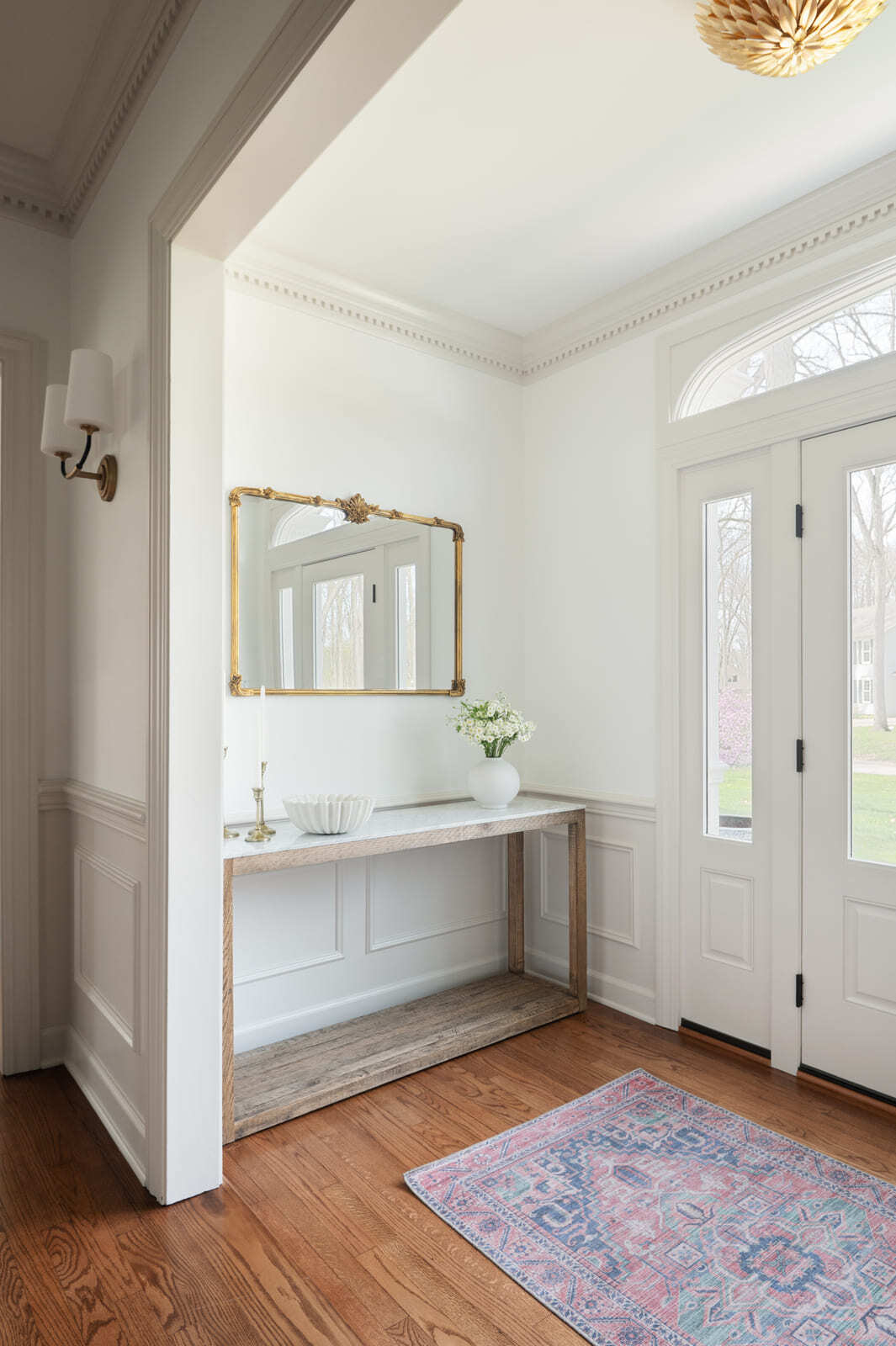 Bright foyer with white trim, gold mirror, wood console, glass front doors, pink rug, and warm wood floors.