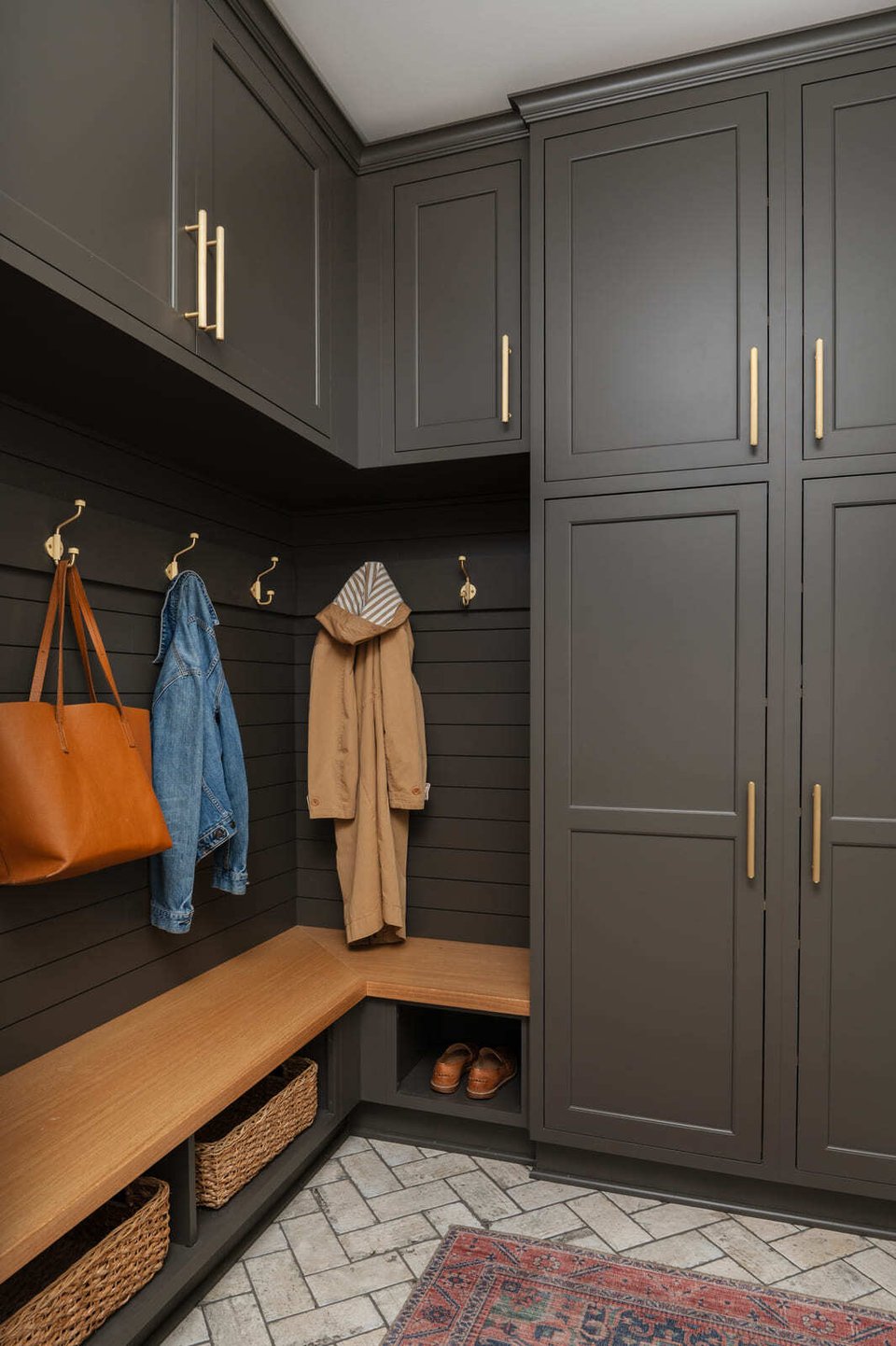 Dark mudroom with tall cabinets, brass hooks, wood bench, baskets, coats, and herringbone tile floor.