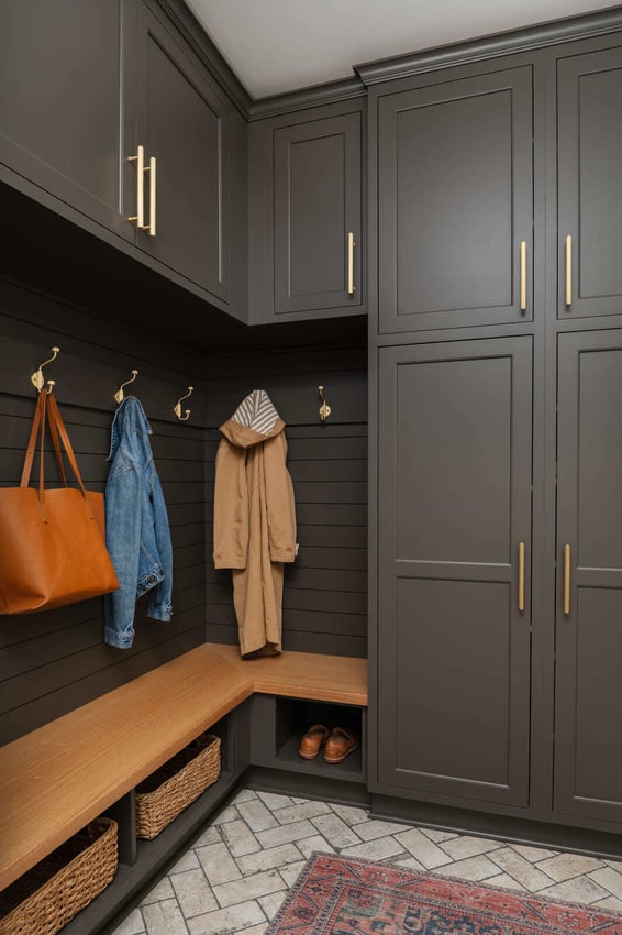 Dark mudroom with tall cabinets, brass hooks, wood bench, baskets, coats, and herringbone tile floor.