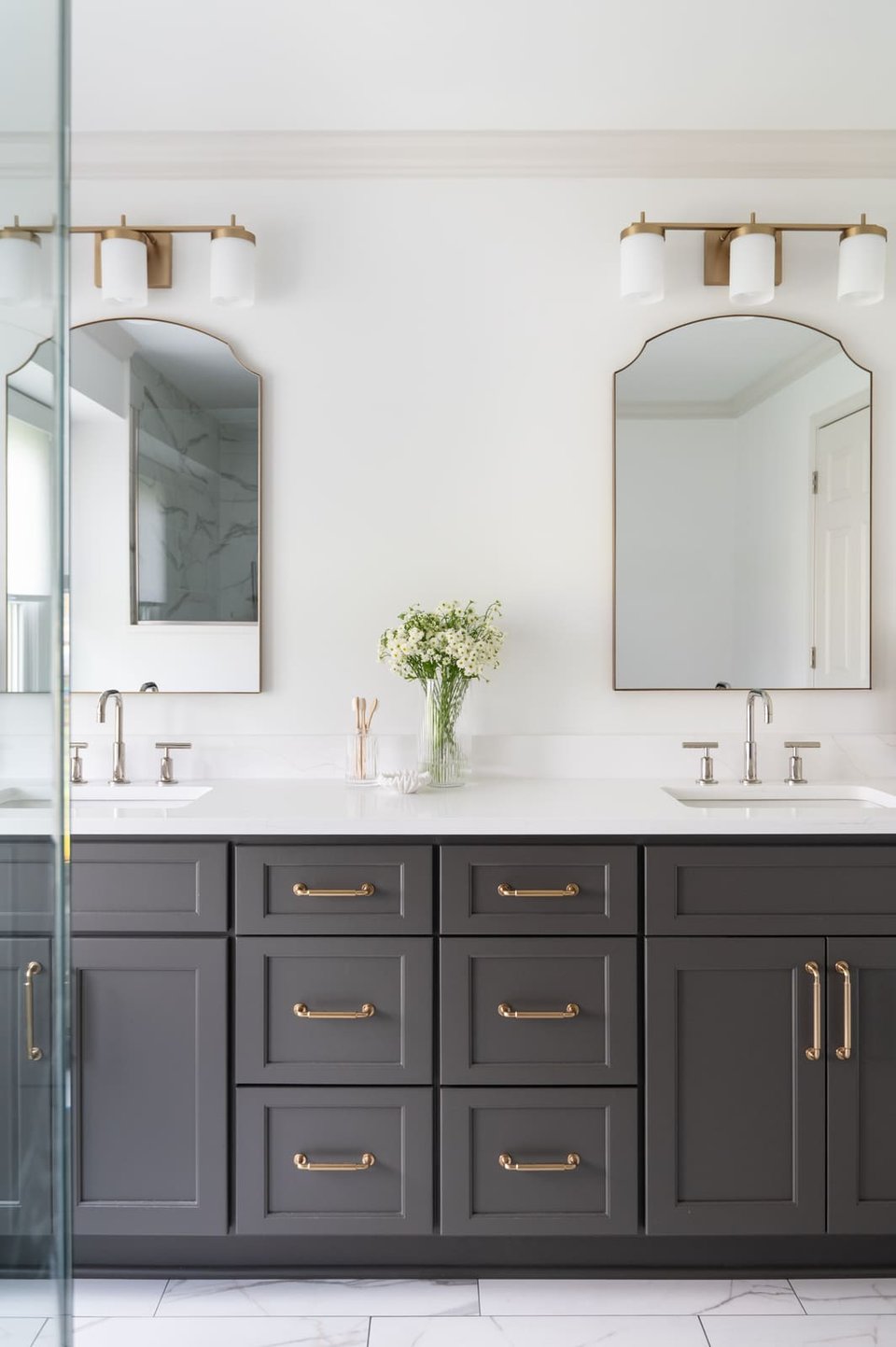 Double bathroom vanity with dark gray cabinets, brass pulls, white counters, arched mirrors, and glass shower.