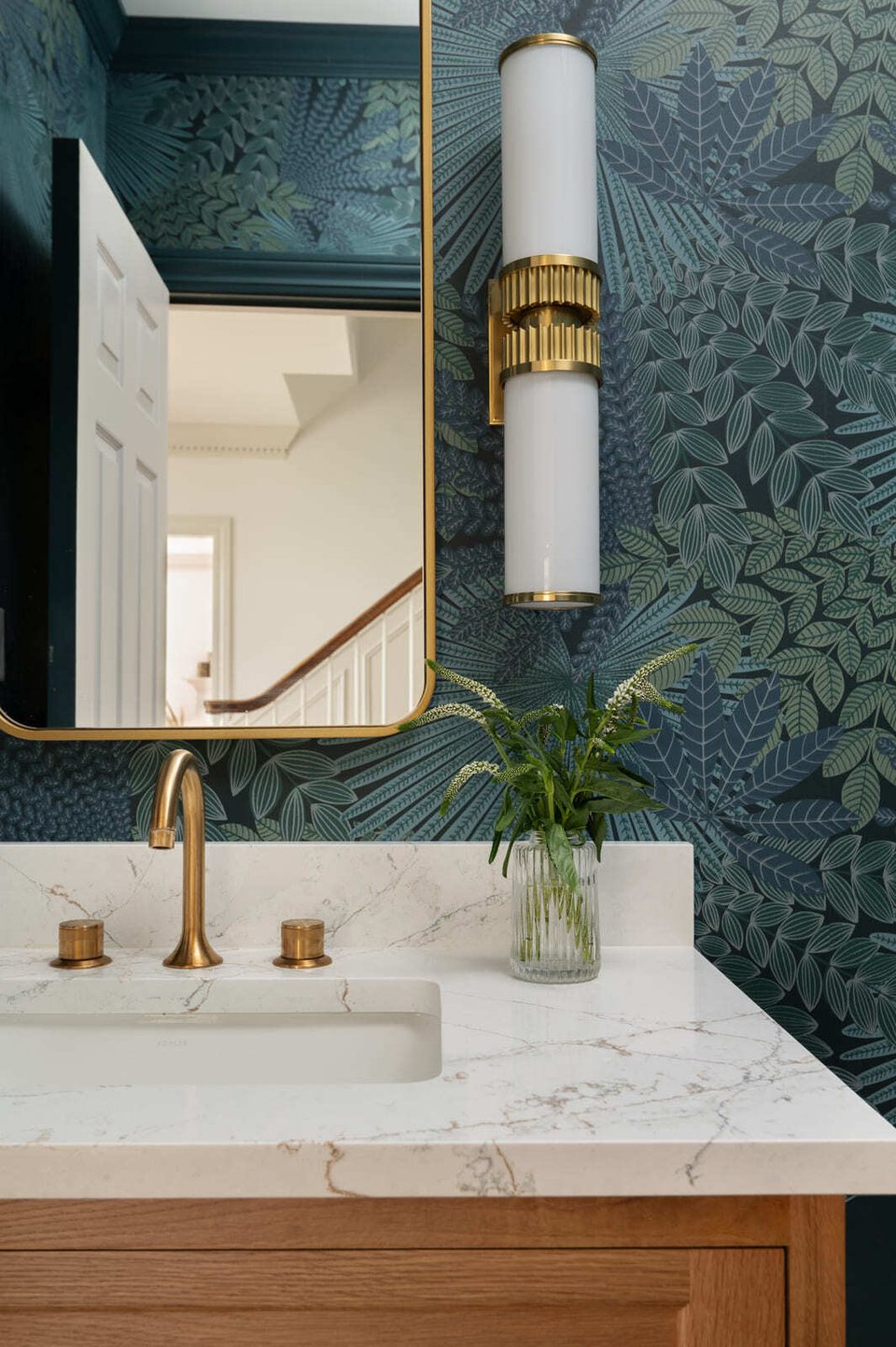 Powder bathroom with botanical wallpaper, brass mirror, white counter, brass faucet, and wall sconce.