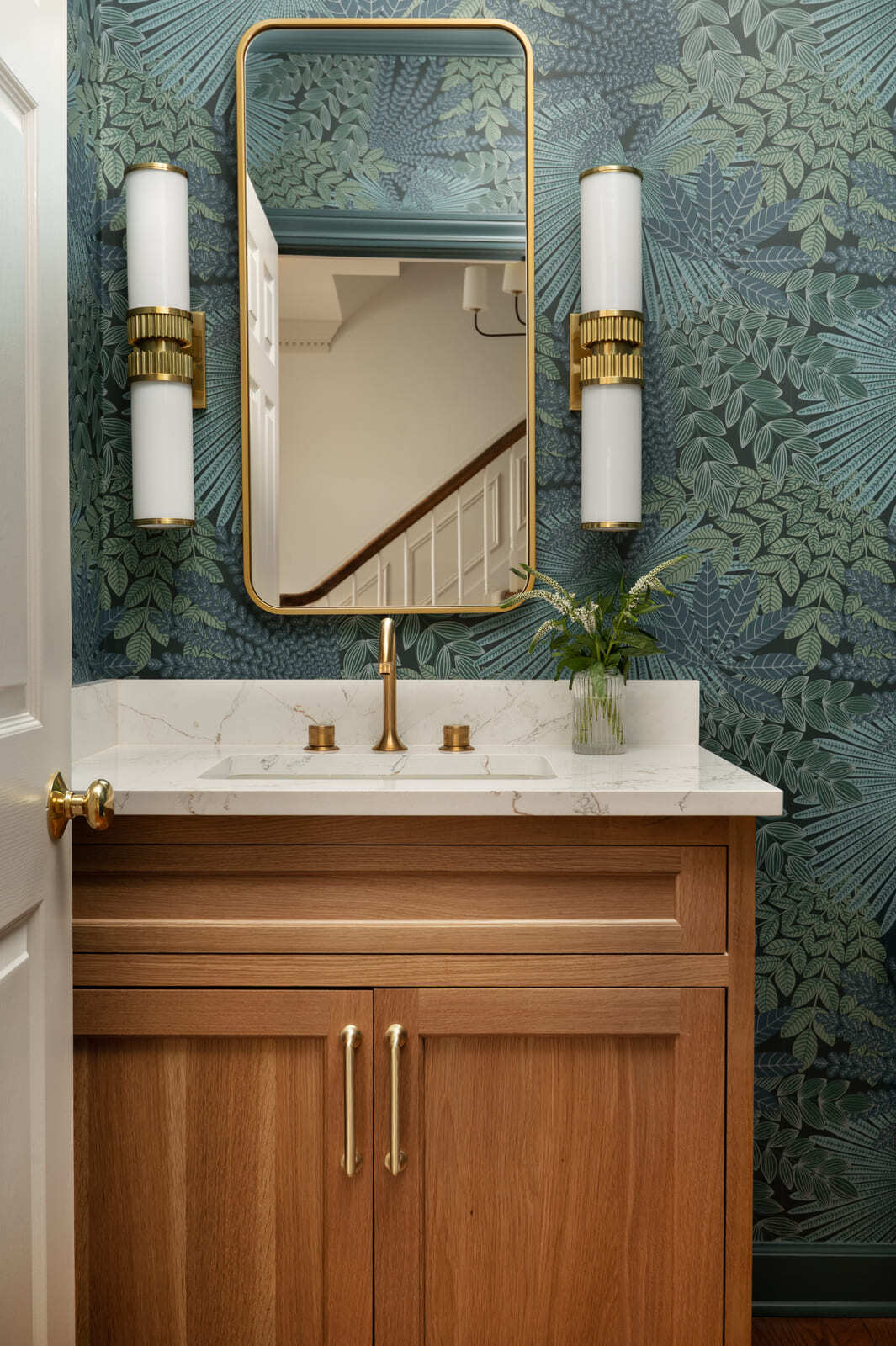 Powder bathroom with botanical wallpaper, wood vanity, white countertop, brass faucet, mirror, and sconces.