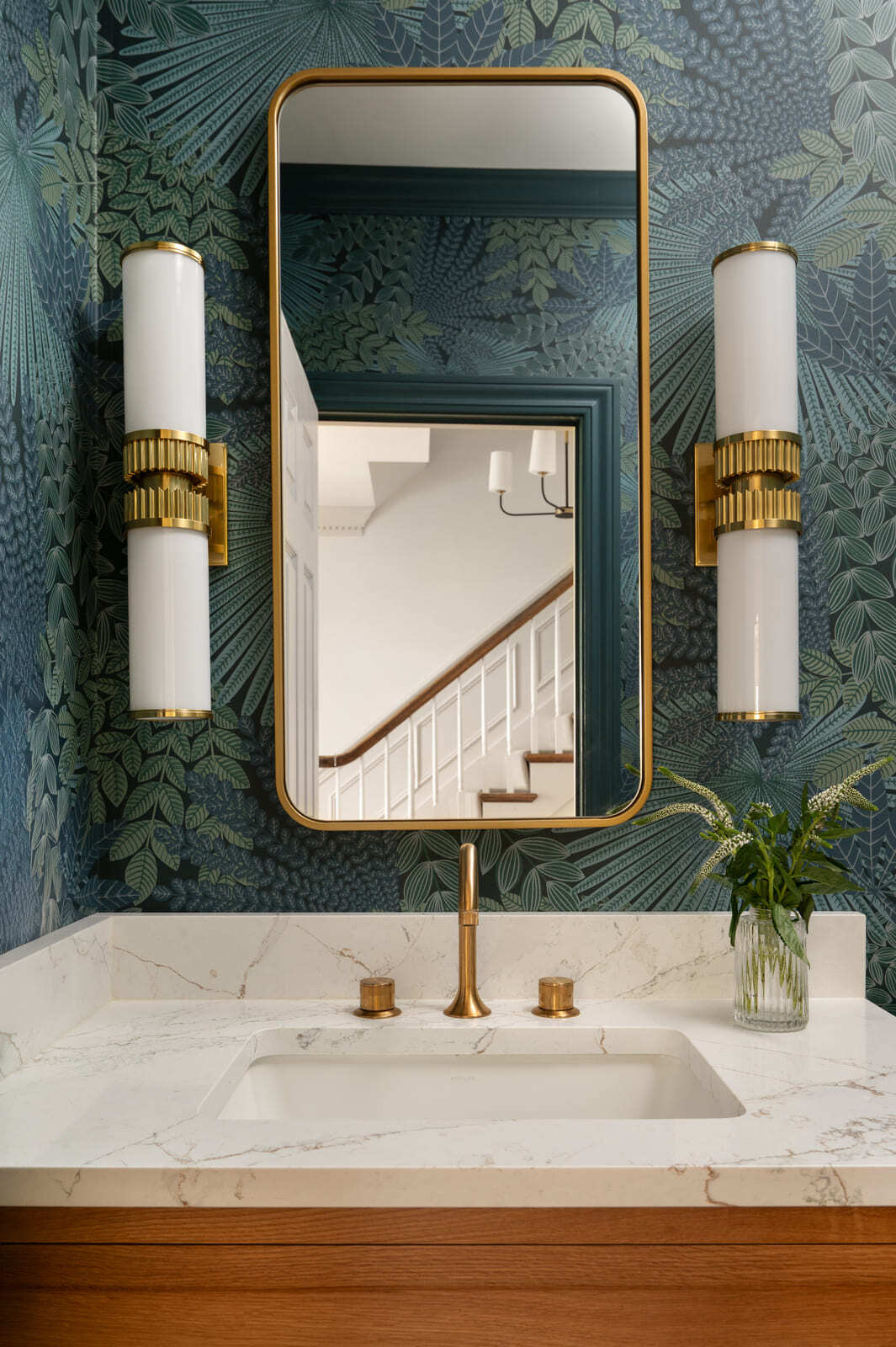 Powder bathroom with green botanical wallpaper, brass mirror, white counter, brass faucet, and white sconces.