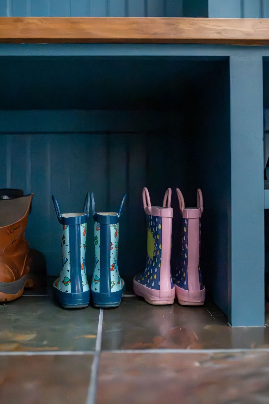 Close-up of kids’ rain boots under blue mudroom bench on slate tile floor located in Indiana